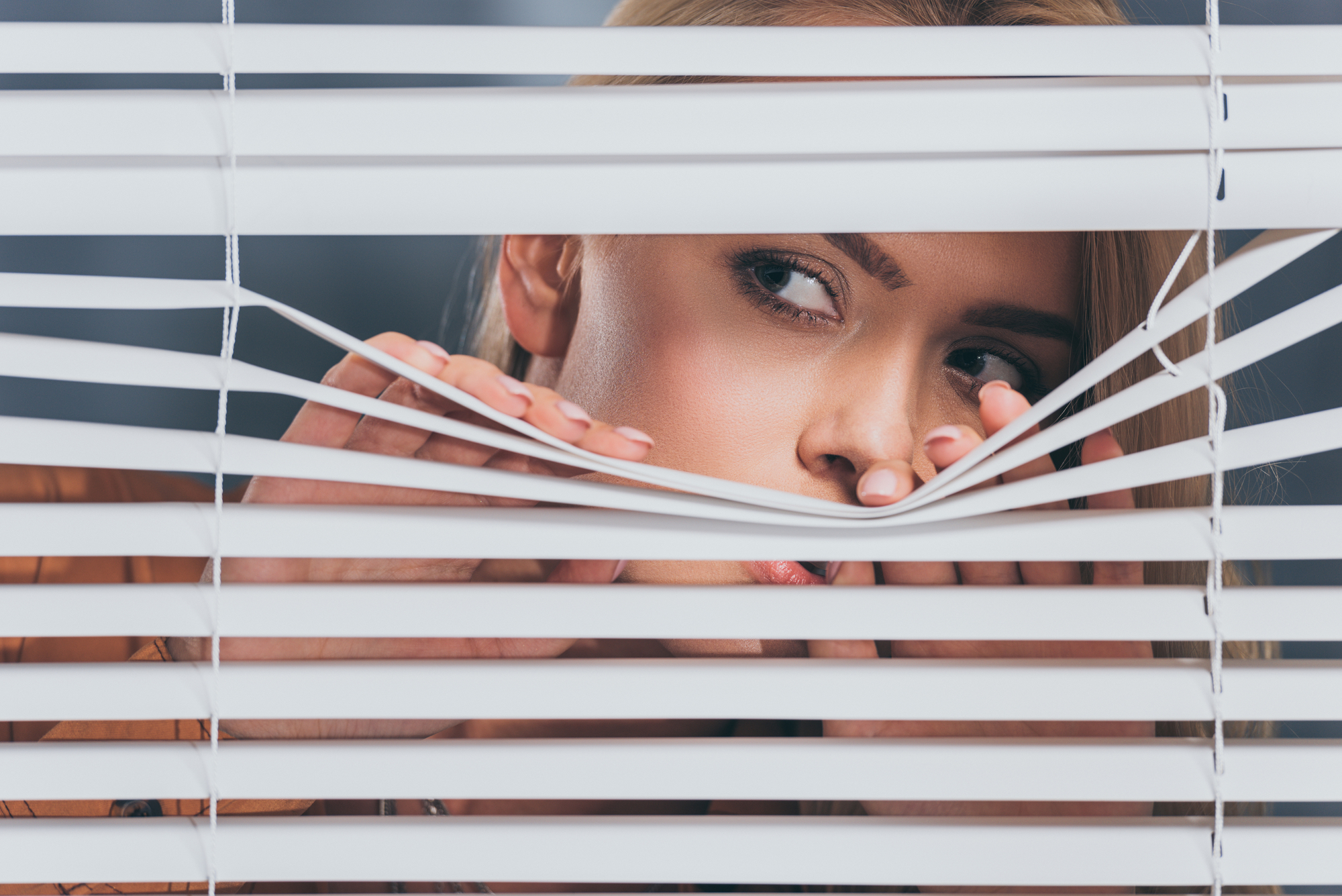 A person peeks through partially opened horizontal blinds. Only their eyes and nose are visible as they stare out with a curious expression. They use their fingers to separate the blinds, creating a narrow gap to look through.