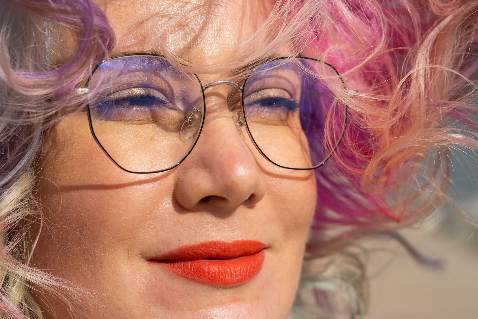 A close-up of a person with colorful hair in shades of purple, pink, and blonde wearing large round glasses. They have vibrant orange lipstick and appear to be outdoors in natural light. Their expression is calm and serene.
