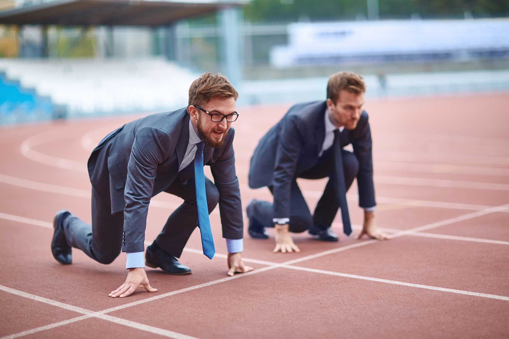 Two men in business suits and ties crouching in starting positions on a running track, ready to race. The background shows a blurred stadium with empty seating. The image symbolizes competition in the business world.