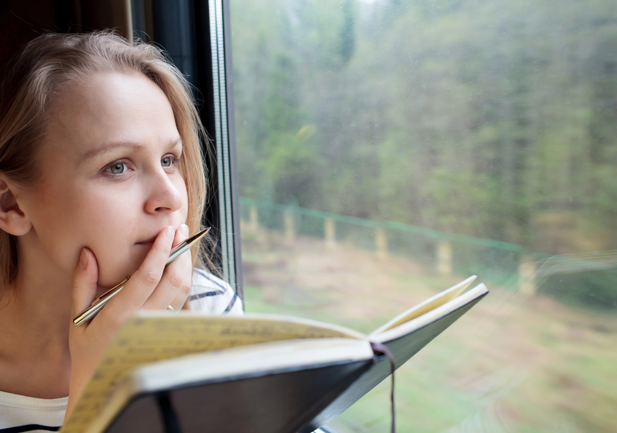 A woman with blonde hair gazes thoughtfully out of a train window, holding a pen to her lips and an open notebook in her lap. The background outside the window shows a blurred view of greenery and trees.
