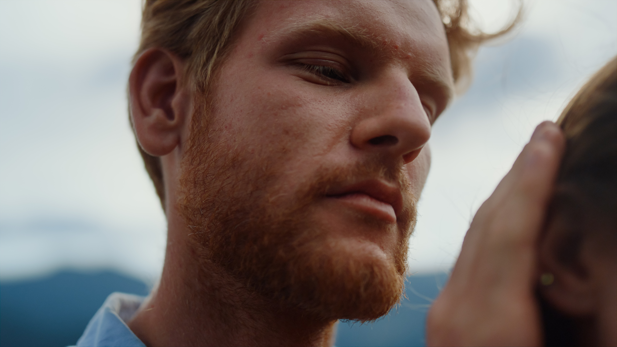 A close-up of a person with short, light brown hair and a beard looking downward, gently touching the side of another person's head. The background is blurred, seemingly an outdoor setting with soft natural light.