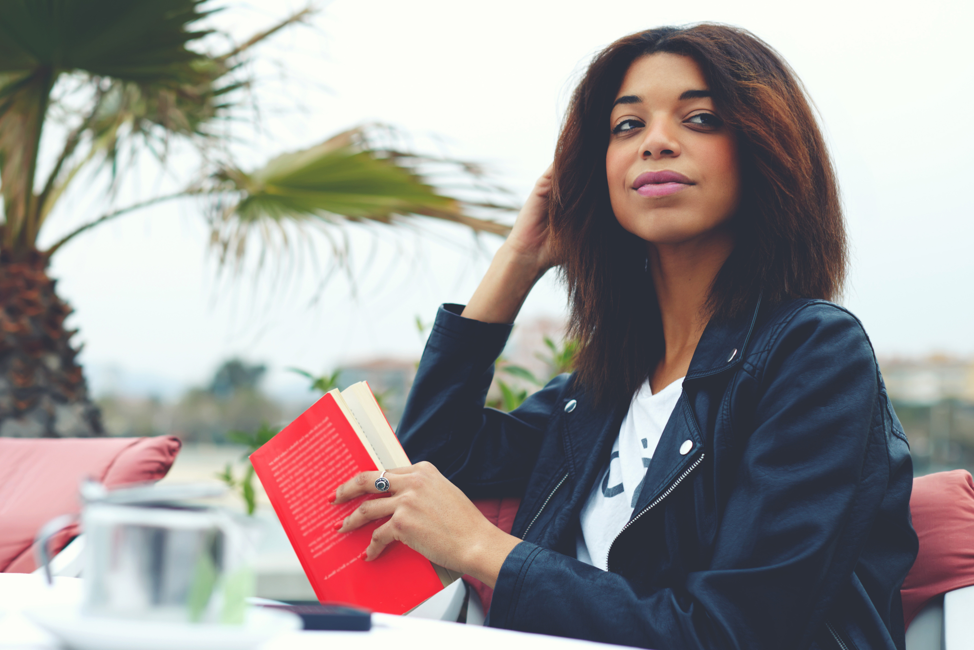 A woman with shoulder-length dark hair, wearing a black leather jacket, is seated outdoors at a table with a red book in her hand. She looks slightly to her right, with palm trees in the background, and a teapot and cup on the table in front of her.