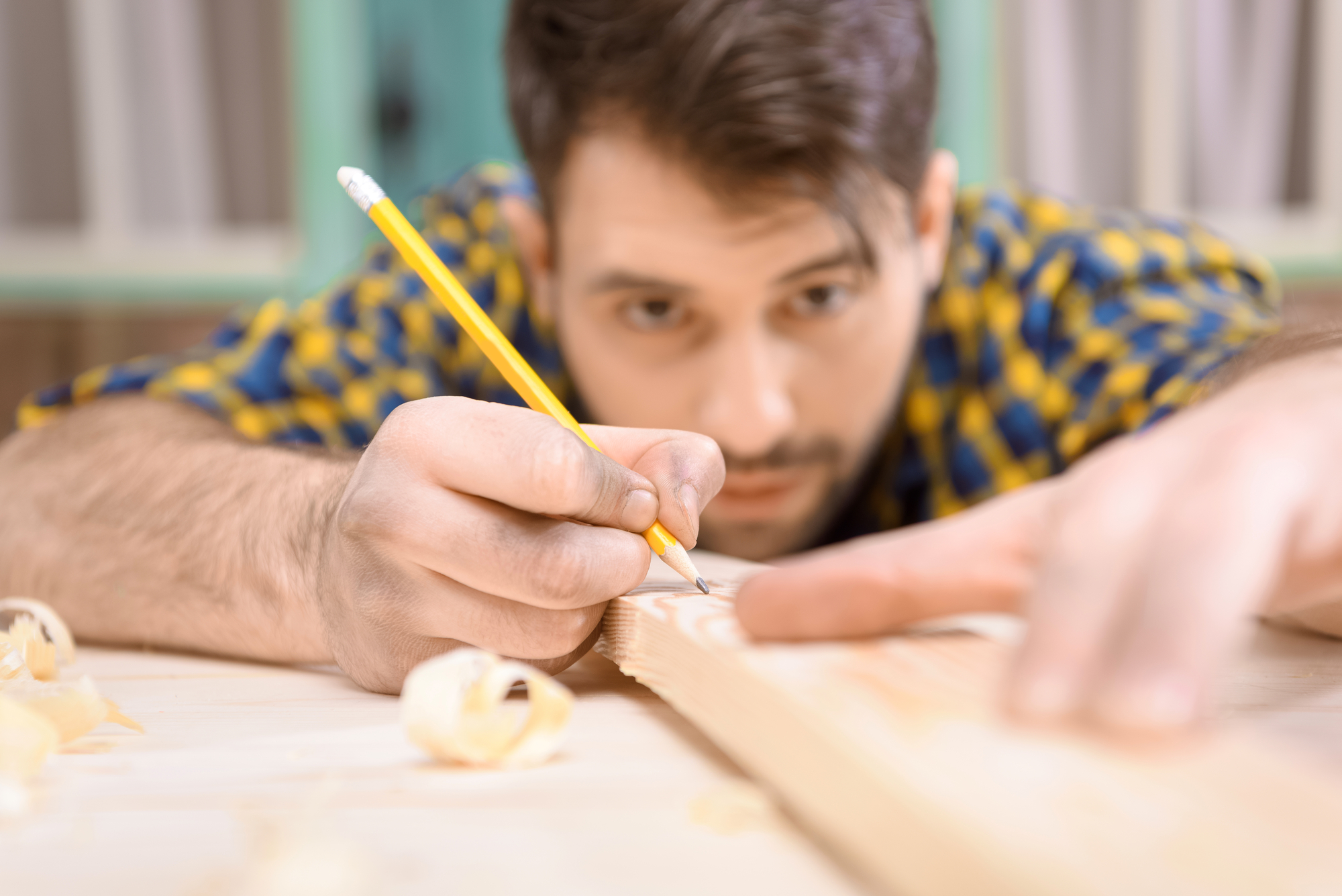 A man in a plaid shirt is carefully marking a piece of wood with a pencil. He is focused on his work, with wood shavings scattered around the table. The background is slightly blurred.