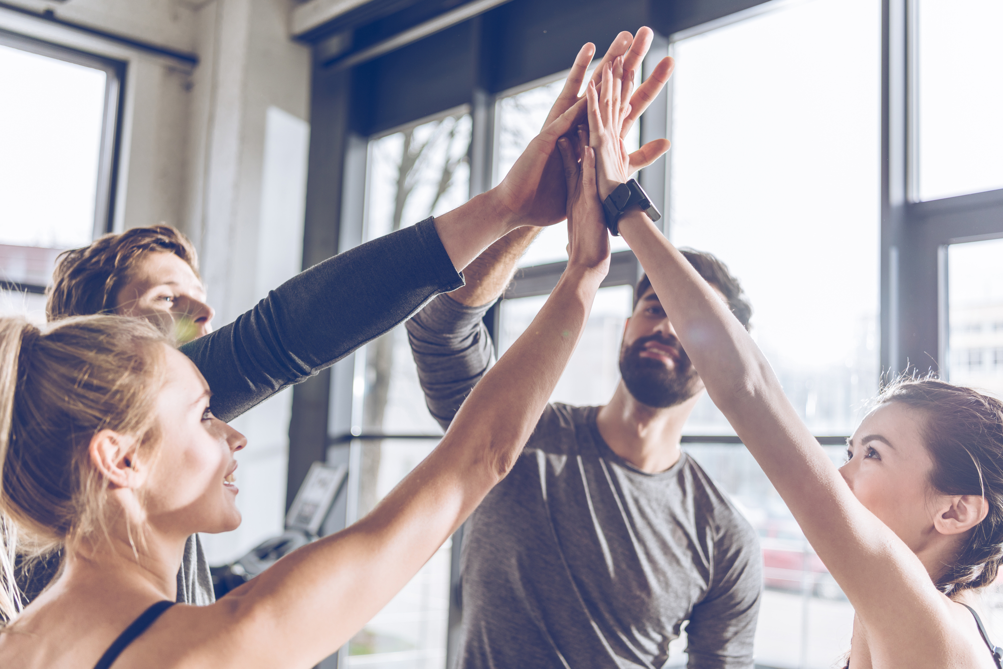 A group of four people in athletic wear are gathered indoors, extending their arms for a group high-five. Sunlight streams through large windows in the background, highlighting their cheerful expressions and sense of teamwork.