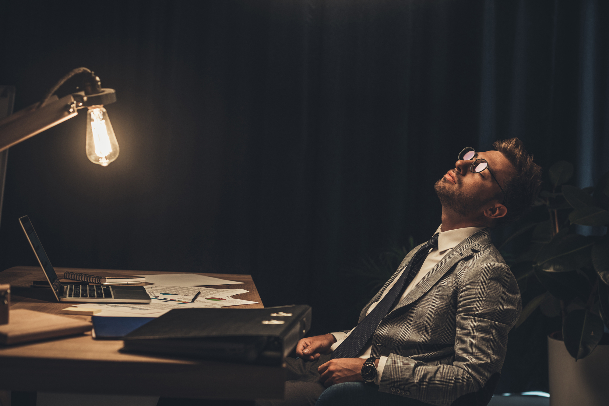 A man in a suit and tie sits at a cluttered desk, leaning back in his chair with his head tilted upward and eyes closed. He appears to be tired or deep in thought. An open laptop and paperwork are visible in front of him, with a desk lamp illuminating the scene.