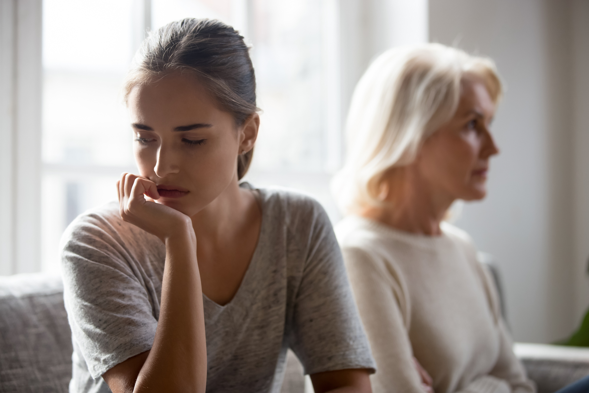 A young woman with her hair tied back, wearing a gray t-shirt, sits in the foreground looking thoughtful and concerned, with her hand resting near her mouth. In the background, an older woman with short, white hair wearing a cream sweater sits turned away. Both appear to be indoors.