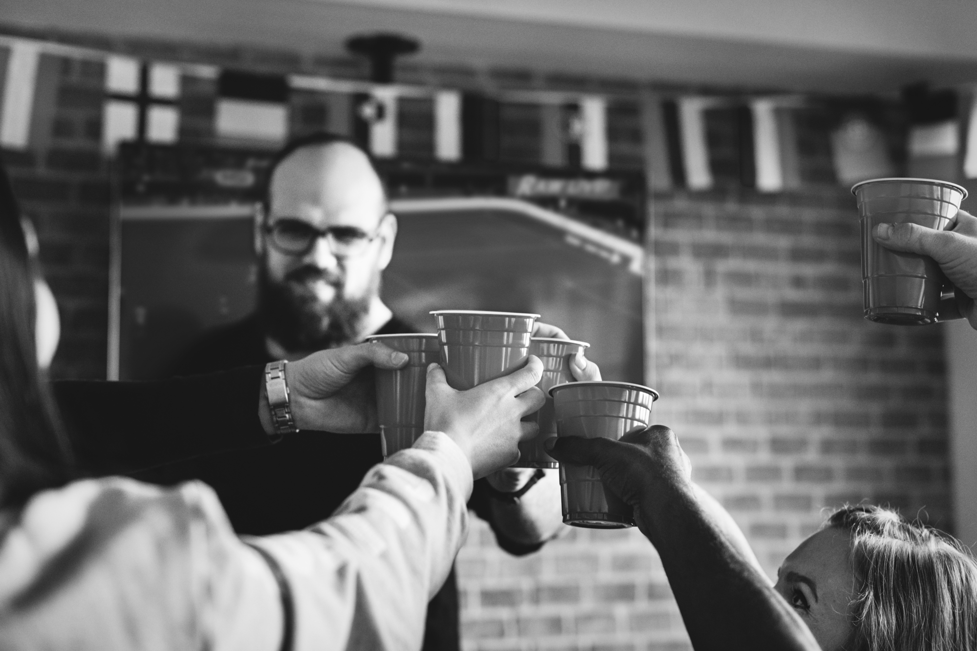 A group of people cheerfully raising plastic cups for a toast in front of a bearded man indoors. They are in a room with a brick wall adorned with hanging flags. The image is in black and white, creating a celebratory and intimate atmosphere.