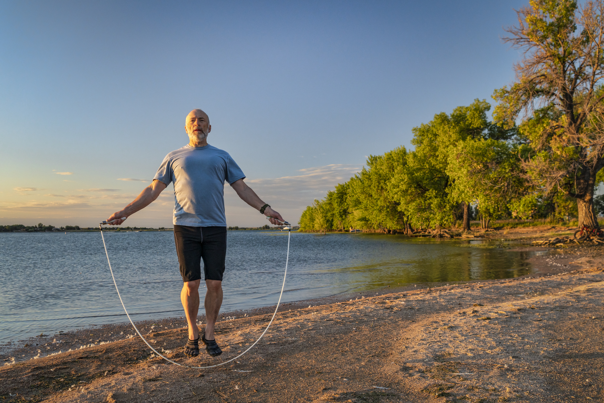 A man with a gray beard, wearing a blue T-shirt and black shorts, jumps rope on a sandy beach by the water. The sky is clear with a few clouds, and lush green trees line the shore in the background. The sun is either rising or setting, casting a warm glow.