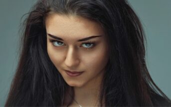 Close-up of a woman with long dark hair and intense blue eyes, looking directly at the camera. She has a neutral expression on her face, with a slightly raised eyebrow and a subtle smile. The background is a soft, medium grey color.