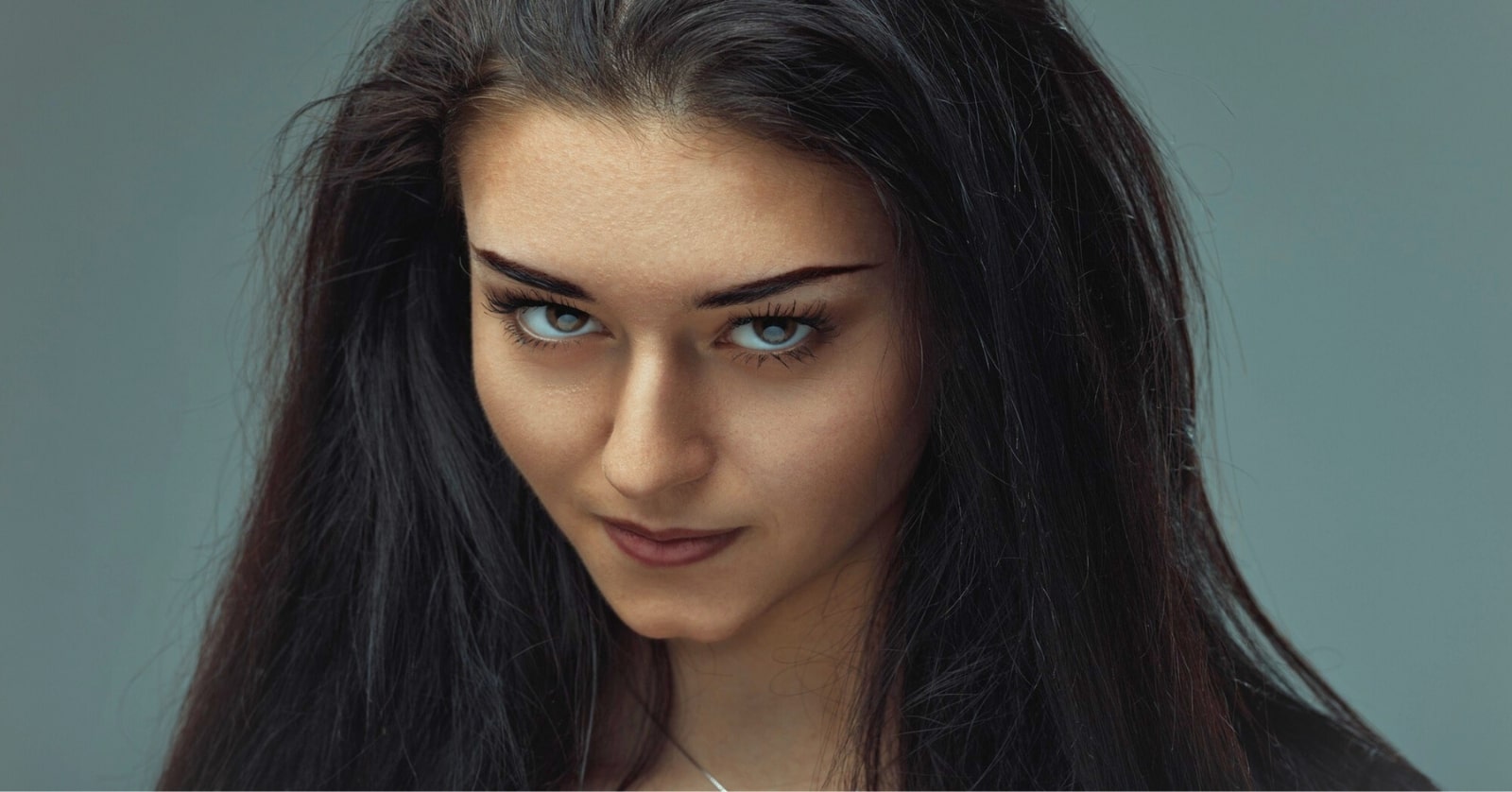 Close-up of a woman with long dark hair and intense blue eyes, looking directly at the camera. She has a neutral expression on her face, with a slightly raised eyebrow and a subtle smile. The background is a soft, medium grey color.