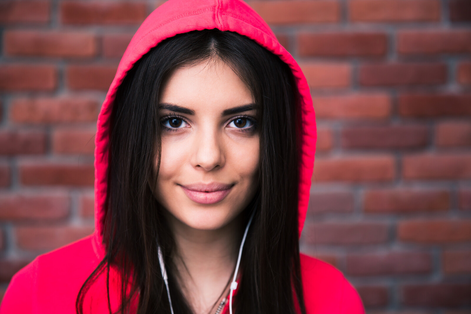 A young woman with long dark hair wearing a bright red hoodie and headphones smiles softly at the camera. She is standing in front of a brick wall background.