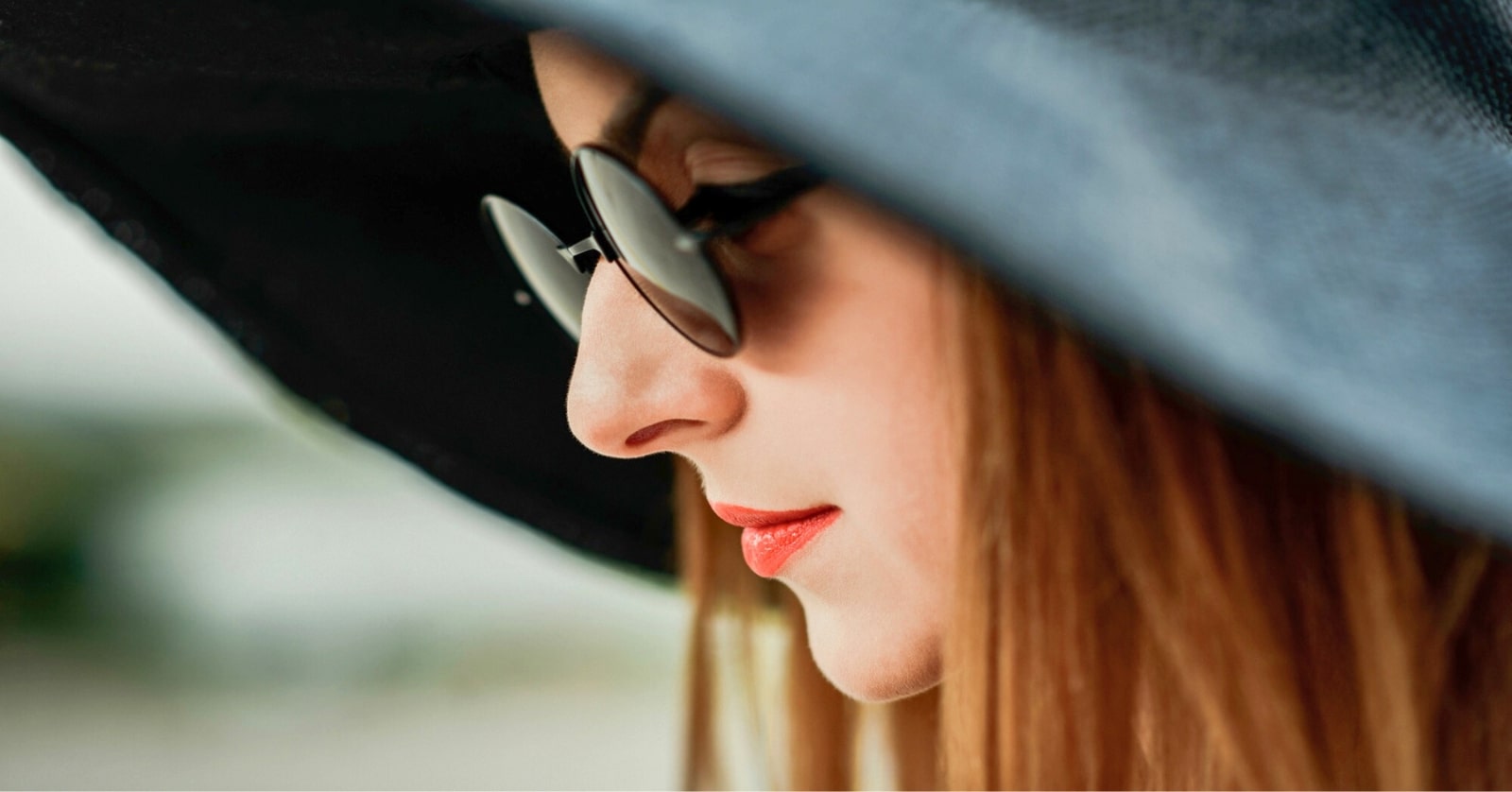 A close-up side profile of a person with long hair, wearing a large dark sun hat and round sunglasses. The individual has vibrant red lipstick and appears to be looking downward, with a blurred background.