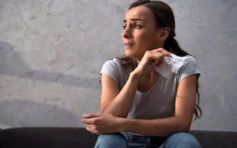 A woman with long hair sits on a couch with a worried expression, holding a tissue in her hand. She is wearing a gray T-shirt and jeans. The background features a plain, light-colored wall.