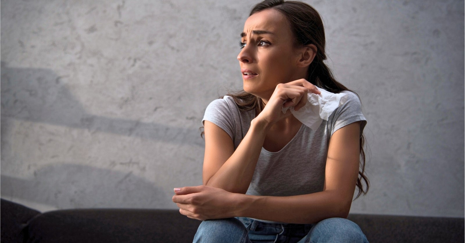 A woman with long hair sits on a couch with a worried expression, holding a tissue in her hand. She is wearing a gray T-shirt and jeans. The background features a plain, light-colored wall.