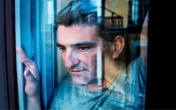 A man with graying hair is looking pensively out of a window. His reflection is visible in the glass, and his hand rests against the window frame. He is wearing a gray shirt, and the scene appears contemplative and introspective.