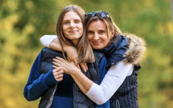 Two smiling women outdoors in a forest setting. The older woman, in a vest with a fur-trimmed hood and sunglasses on her head, hugs the younger woman, who is also wearing a vest and has long brown hair. They both look at the camera with smiles.