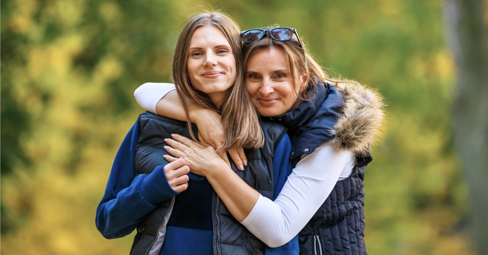 Two smiling women outdoors in a forest setting. The older woman, in a vest with a fur-trimmed hood and sunglasses on her head, hugs the younger woman, who is also wearing a vest and has long brown hair. They both look at the camera with smiles.