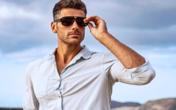 A man with short, styled hair is standing outdoors against a cloudy sky. He is wearing a light blue shirt and dark sunglasses, with one arm raised to adjust the sunglasses. The background includes distant mountainous terrain.