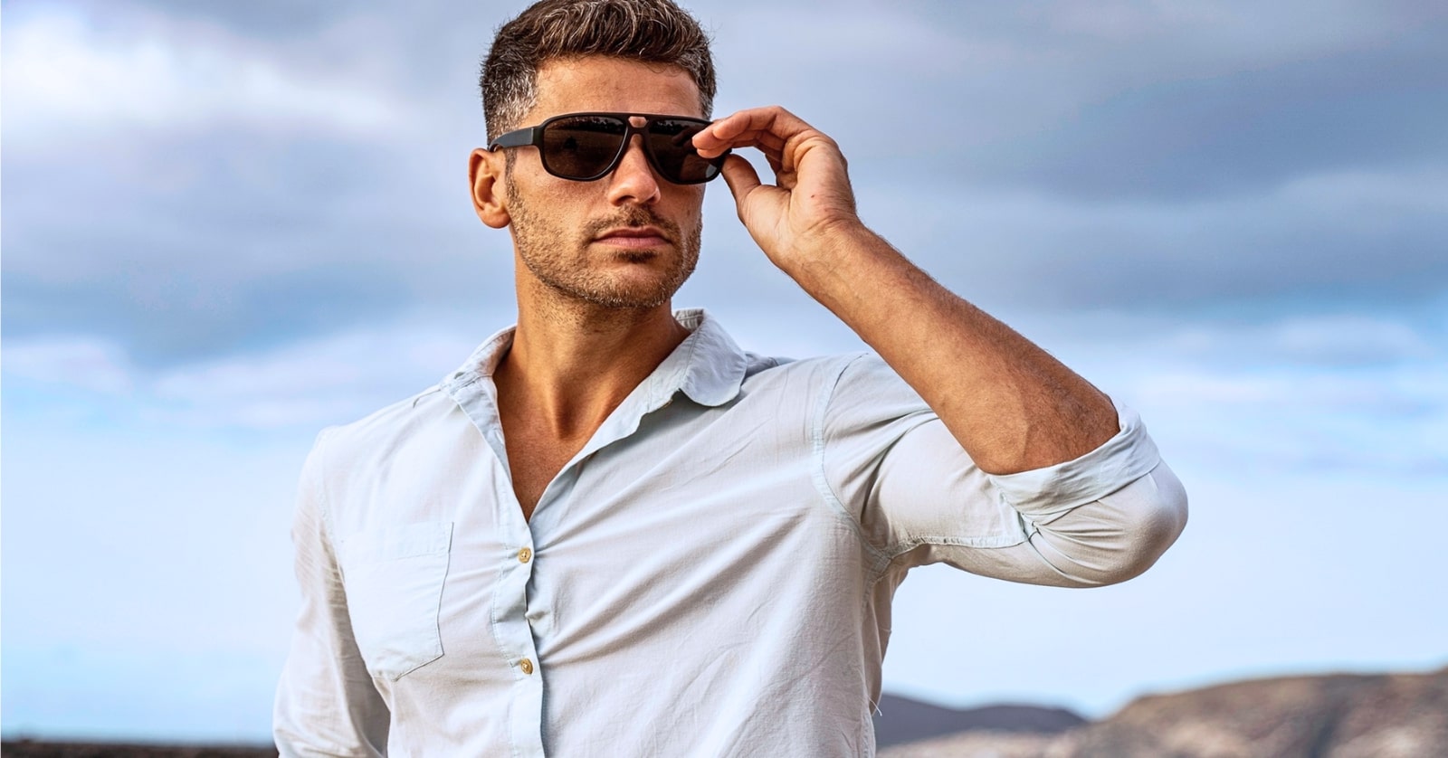 A man with short, styled hair is standing outdoors against a cloudy sky. He is wearing a light blue shirt and dark sunglasses, with one arm raised to adjust the sunglasses. The background includes distant mountainous terrain.