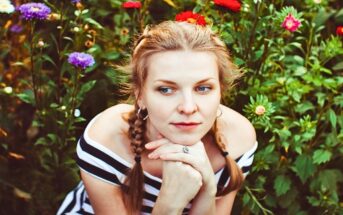 A woman with braided hair, wearing a black and white striped off-shoulder top, rests her chin on her hands while sitting amidst a garden of colorful flowers, including red, purple, and pink blooms. She gazes thoughtfully into the distance.