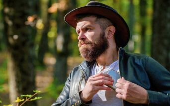 a rugged looking man with beard and hat walks through the forest and puts a metal flask back into his jacket pocket