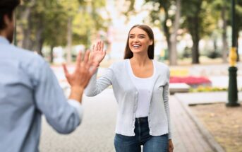 A woman with long brown hair, wearing a white shirt and light gray cardigan, smiles as she walks on a tree-lined path and high-fives a person in a light blue shirt. The background shows greenery and blurred park scenery.