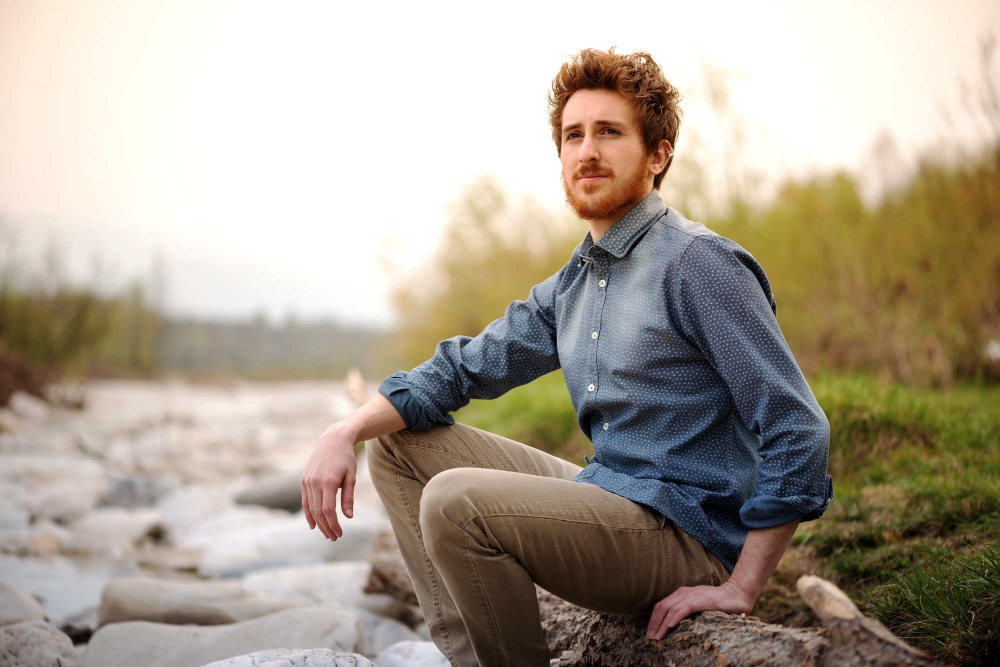 A man with curly hair and a beard is sitting on a large rock beside a riverbank. He is wearing a blue buttoned-down shirt with rolled-up sleeves and khaki pants. The background features trees and a cloudy sky. The scene exudes a calm, contemplative mood.