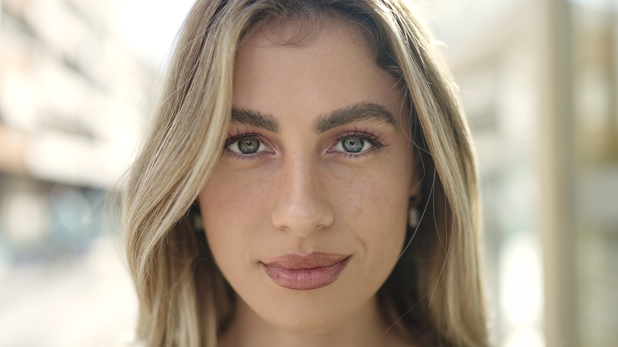 Close-up portrait of a young woman with light brown hair and blue eyes. She is looking directly at the camera with a calm expression. The background is blurred, suggesting an outdoor setting.