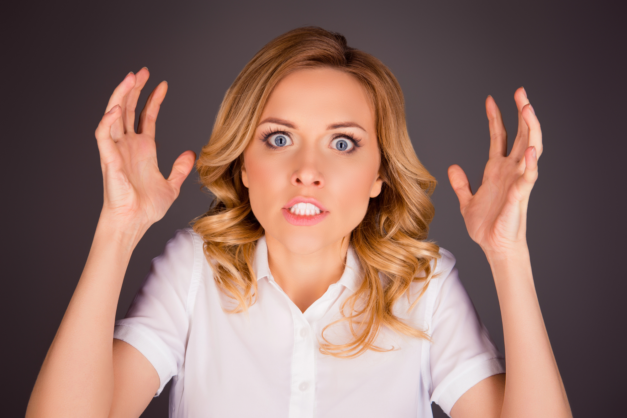 A woman with blonde hair, wearing a white shirt, has an angry expression. Her eyes are wide open, mouth slightly open showing teeth, and her hands are raised with fingers spread apart. The background is dark, making her face and gestures more prominent.