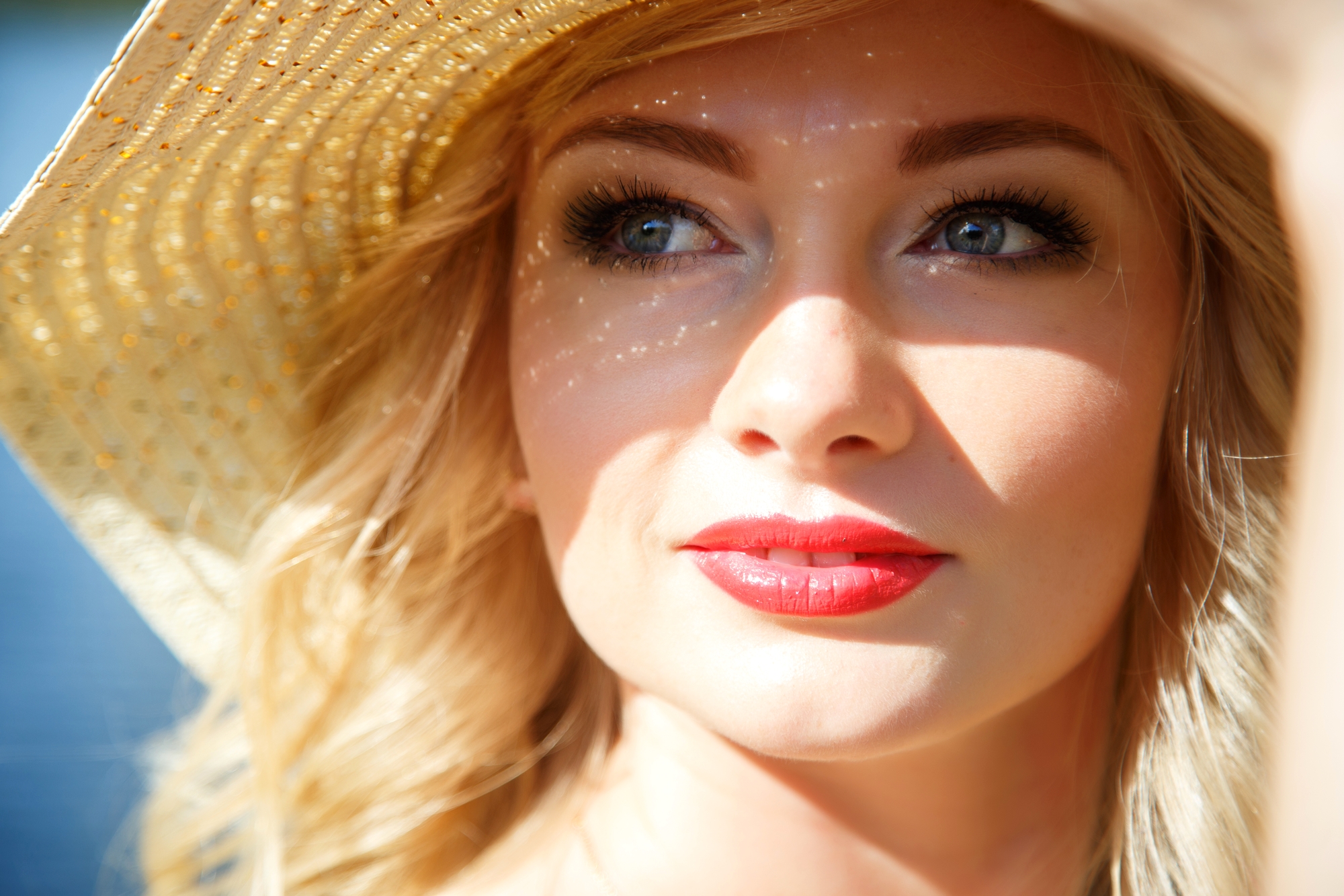 A close-up of a person with long blonde hair wearing a wide-brimmed hat. The hat casts a pattern of dappled sunlight on their face. They have blue eyes, prominent eyelashes, and red lipstick. The background is blurred and out of focus.