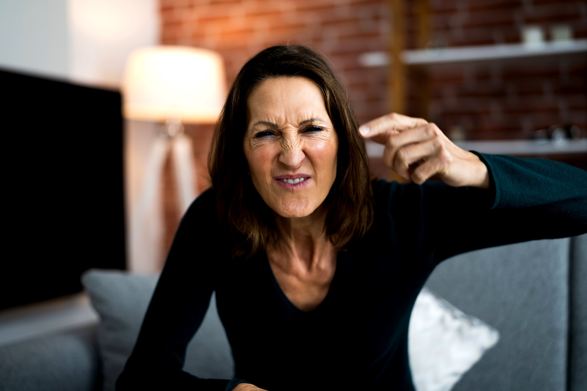 A woman with shoulder-length brown hair is seated on a grey couch, making a frustrated or angry facial expression while pointing her finger forward. The background includes a lit floor lamp and a brick wall.