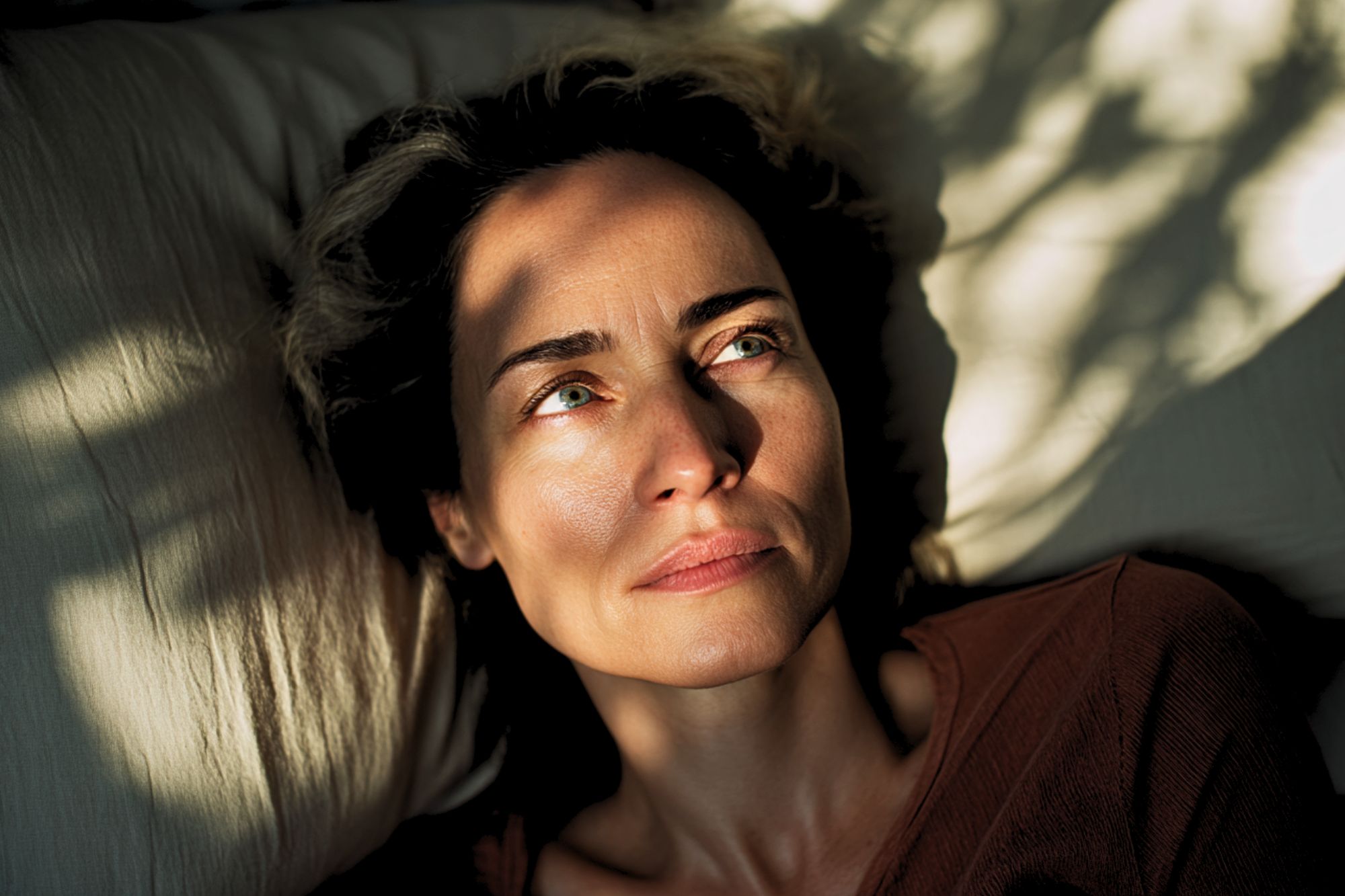 A woman with green eyes lies on a pillow, looking upwards, with dappled sunlight and shadows across her face.
