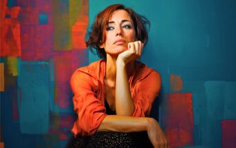 A woman with short brown hair in an orange shirt rests her chin on her hand, looking thoughtful, in front of a blue wall with colorful abstract shapes.