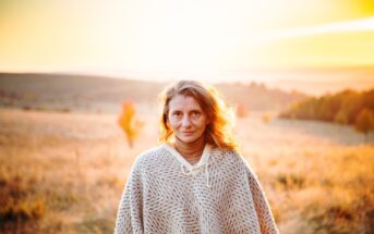 A person with wavy hair stands in a field at sunset, wearing a beige knitted sweater. The background features an autumn landscape with rolling hills and trees displaying fall colors, radiating a warm, golden glow.