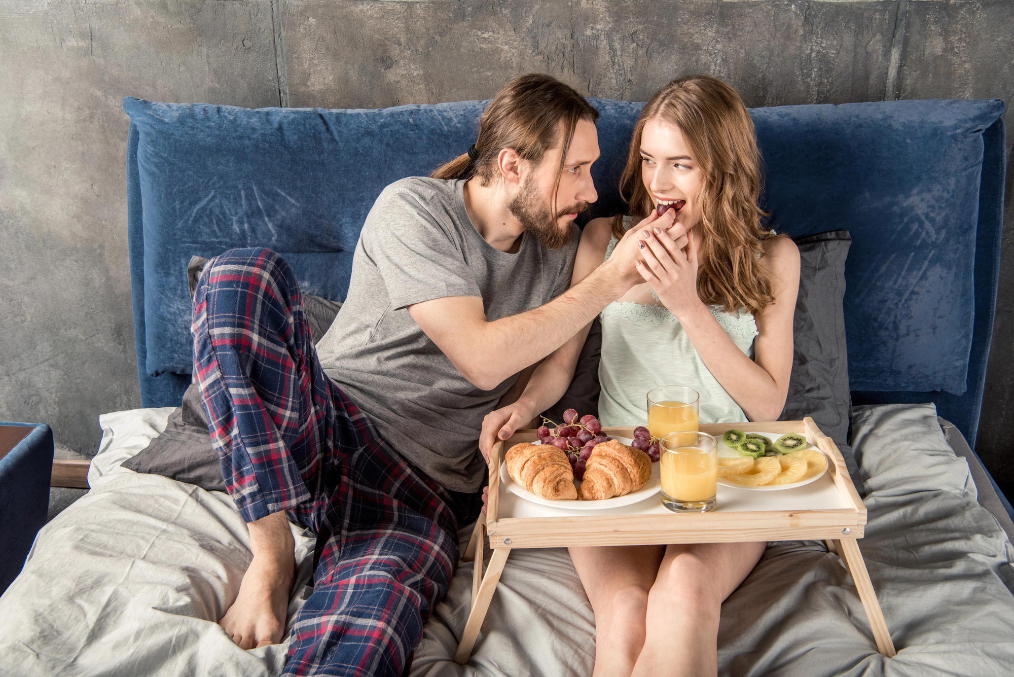 A man and woman are sitting on a bed with a blue headboard. The man, dressed in a grey t-shirt and plaid pants, is feeding the woman, who is wearing a light green camisole. A breakfast tray with croissants, fruits, and glasses of juice is placed on the bed.