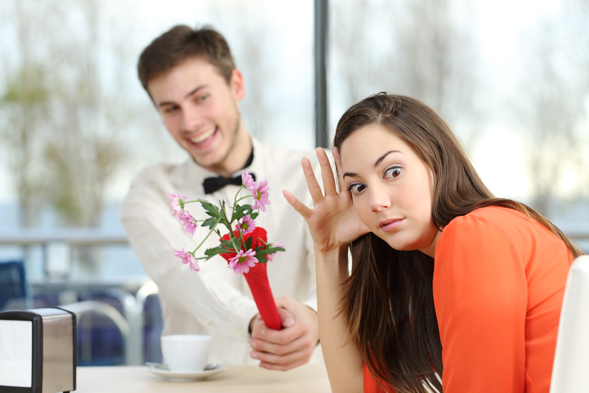 A man smiling and offering a bouquet of flowers to a woman who appears uninterested or uncomfortable. They are seated at a table in a bright setting with a window view. The woman is leaning back with a skeptical expression.