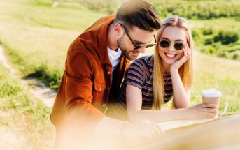 A young couple outdoors, standing beside a car. The man, in a brown jacket and sunglasses, looks at a map, while the woman, with long blonde hair, sunglasses, and a striped shirt, smiles and holds a takeaway coffee cup in the countryside.