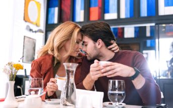 A couple sits closely at a café table, lovingly touching foreheads. Both hold coffee cups, and the table is set with water glasses, a carafe, and a small vase with flowers. The background features modern, colorful abstract art on the wall.