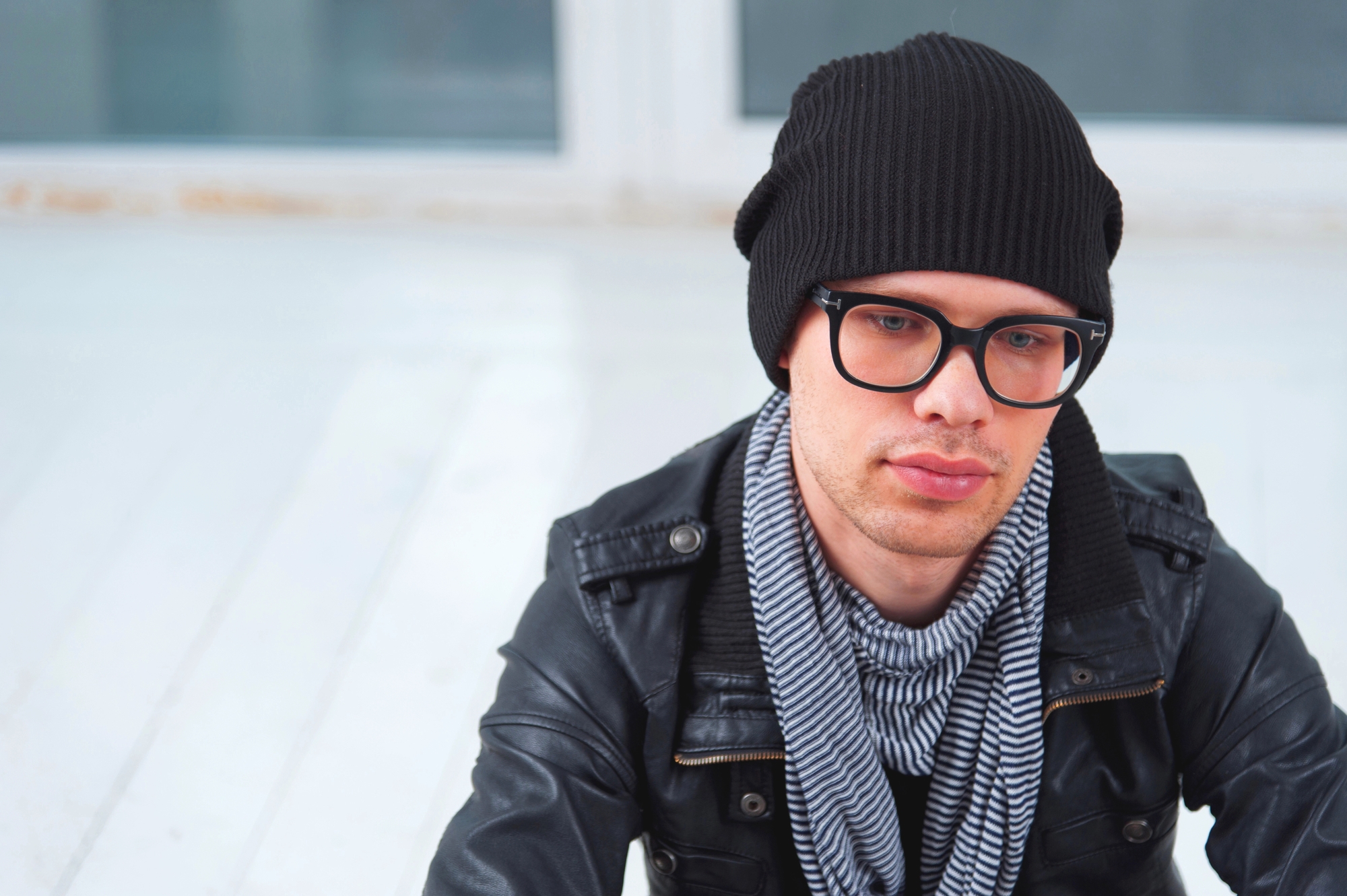 A person with glasses, wearing a black beanie, black leather jacket, and a striped scarf, is sitting in an outdoor setting with a light-colored wooden floor and blurred background. The person appears to be looking off to the side with a neutral expression.