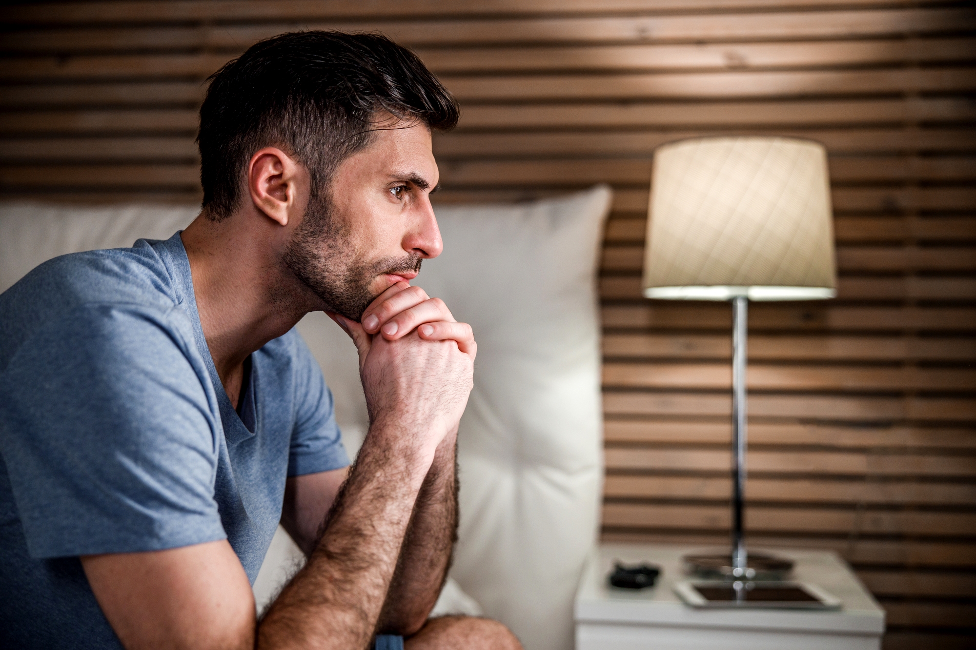 A man with short dark hair and a beard, wearing a gray t-shirt, is sitting on the edge of a bed with his hands clasped under his chin, looking forward thoughtfully. A bedside table with a lamp and a phone is beside him. The background has wooden paneling.