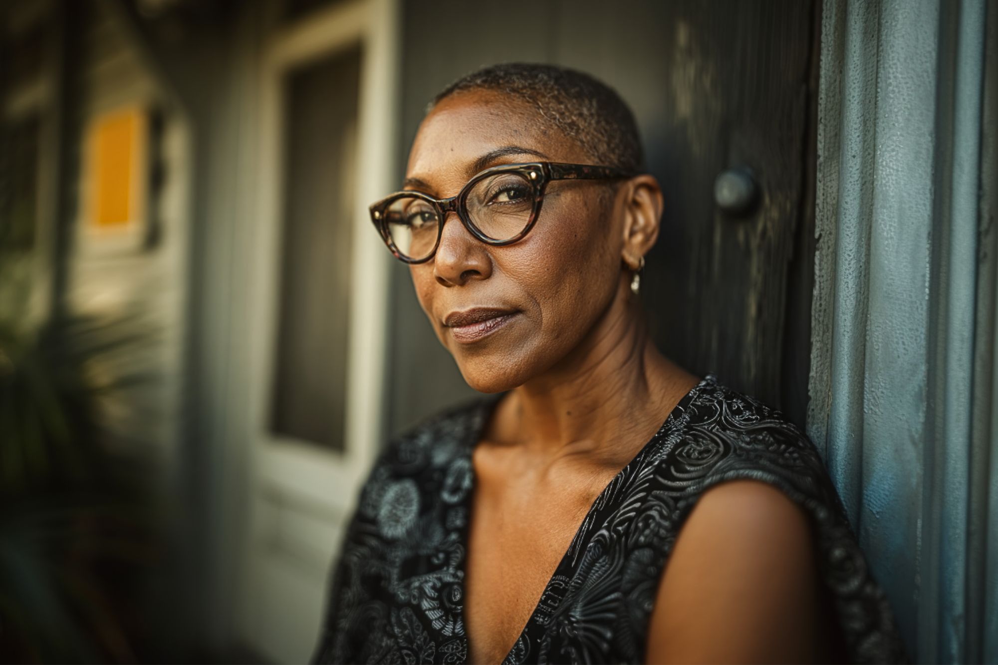 A woman with short hair and glasses, wearing a patterned top, looks confidently at the camera while leaning against a wall outside a building. The background is softly blurred.