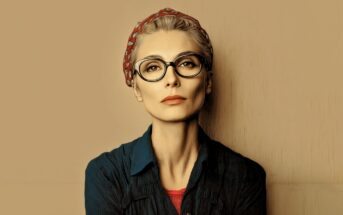 A woman with short blond hair, wearing large black glasses and a red headband, looks forward with a neutral expression. She is dressed in a dark blue shirt over a red top against a beige background.