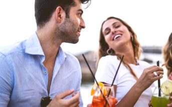 Two people enjoying drinks and smiling in an outdoor setting. The person on the left is wearing a light blue shirt and holding a drink with an orange slice. The person on the right is wearing a white top, holding a drink, and laughing. The background is bright and blurred.