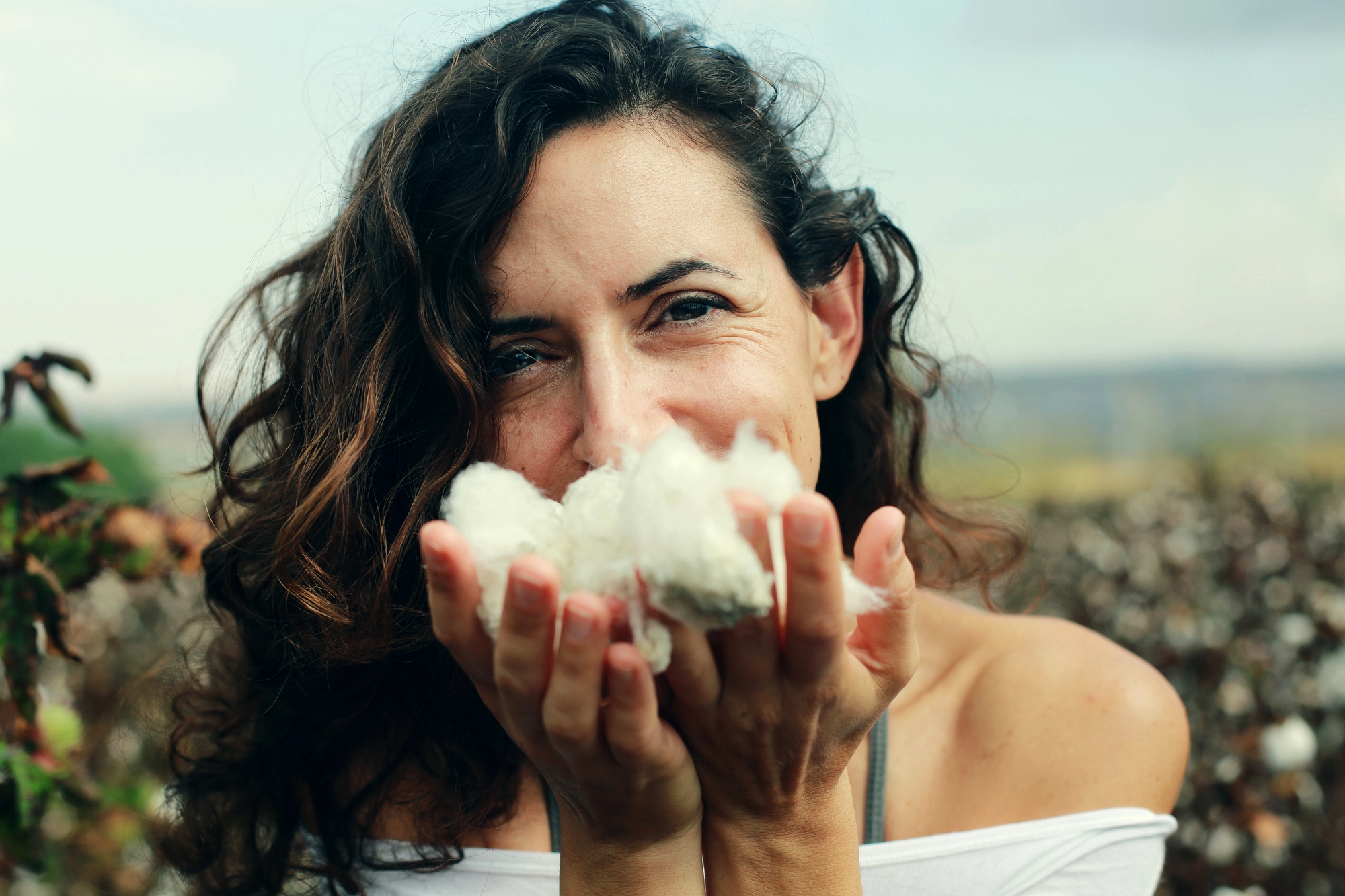 A woman with wavy brown hair holds fluffy white cotton close to her face, smiling gently. She is outdoors in a field, with blurred cotton plants visible in the background, wearing a white off-the-shoulder top.