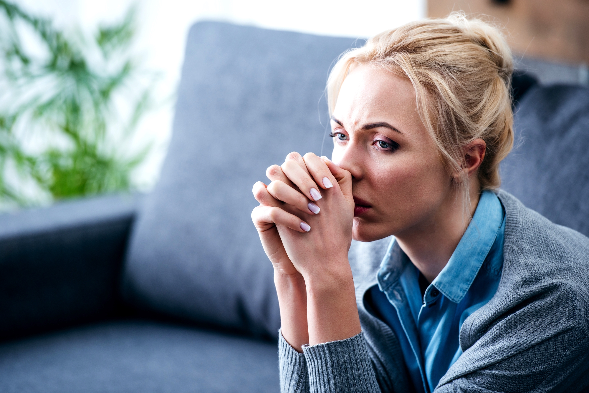 A woman with blonde hair sits on a gray couch, leaning forward with hands clasped near her face, appearing deep in thought or worried. She wears a light blue collared shirt and a gray cardigan. The background is blurred with some greenery visible.