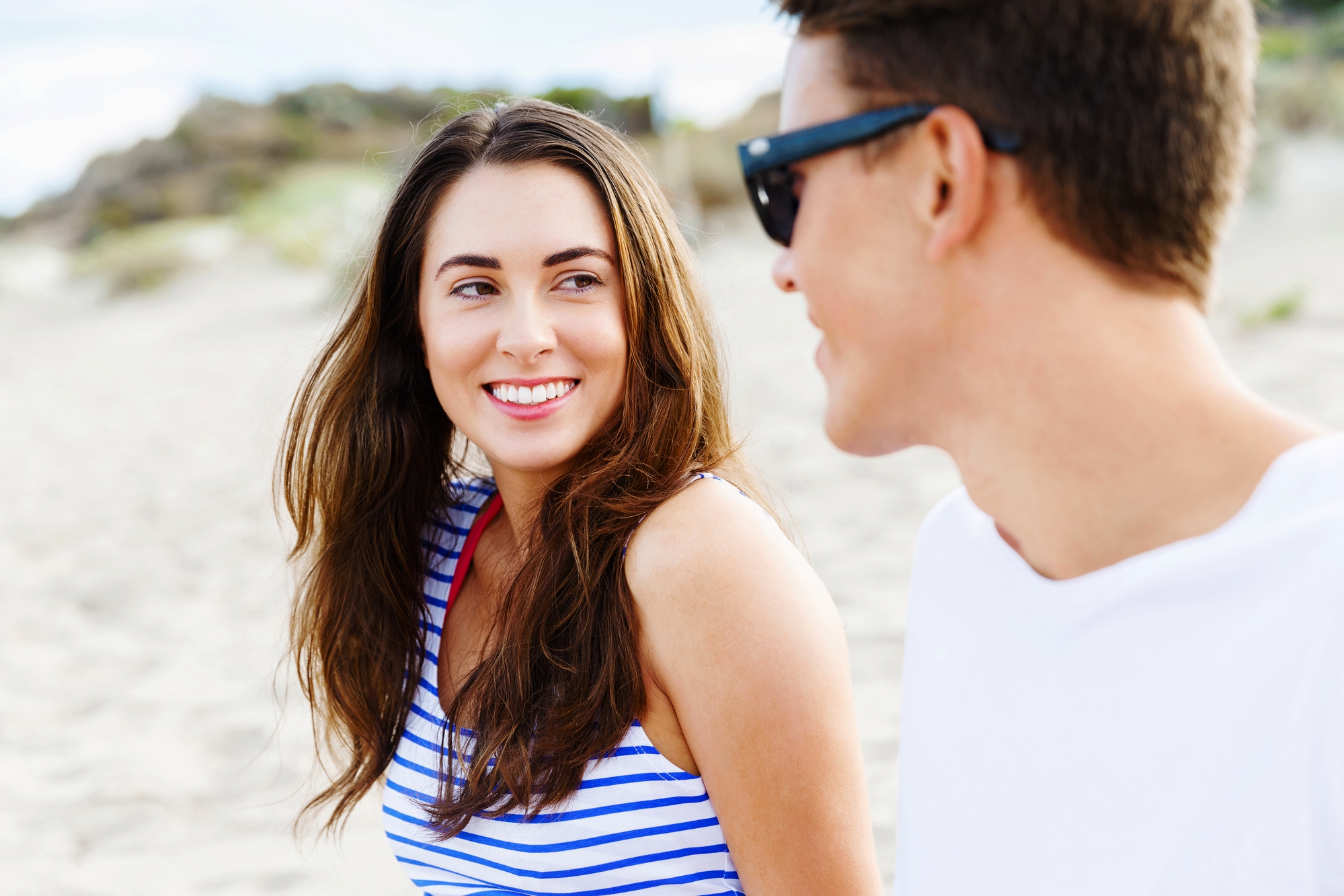 A woman with long brown hair, wearing a striped tank top, smiles at a man with short brown hair and sunglasses. They are sitting together on a sandy beach with blurred greenery in the background. The man is turned towards the woman, appearing to enjoy the moment.