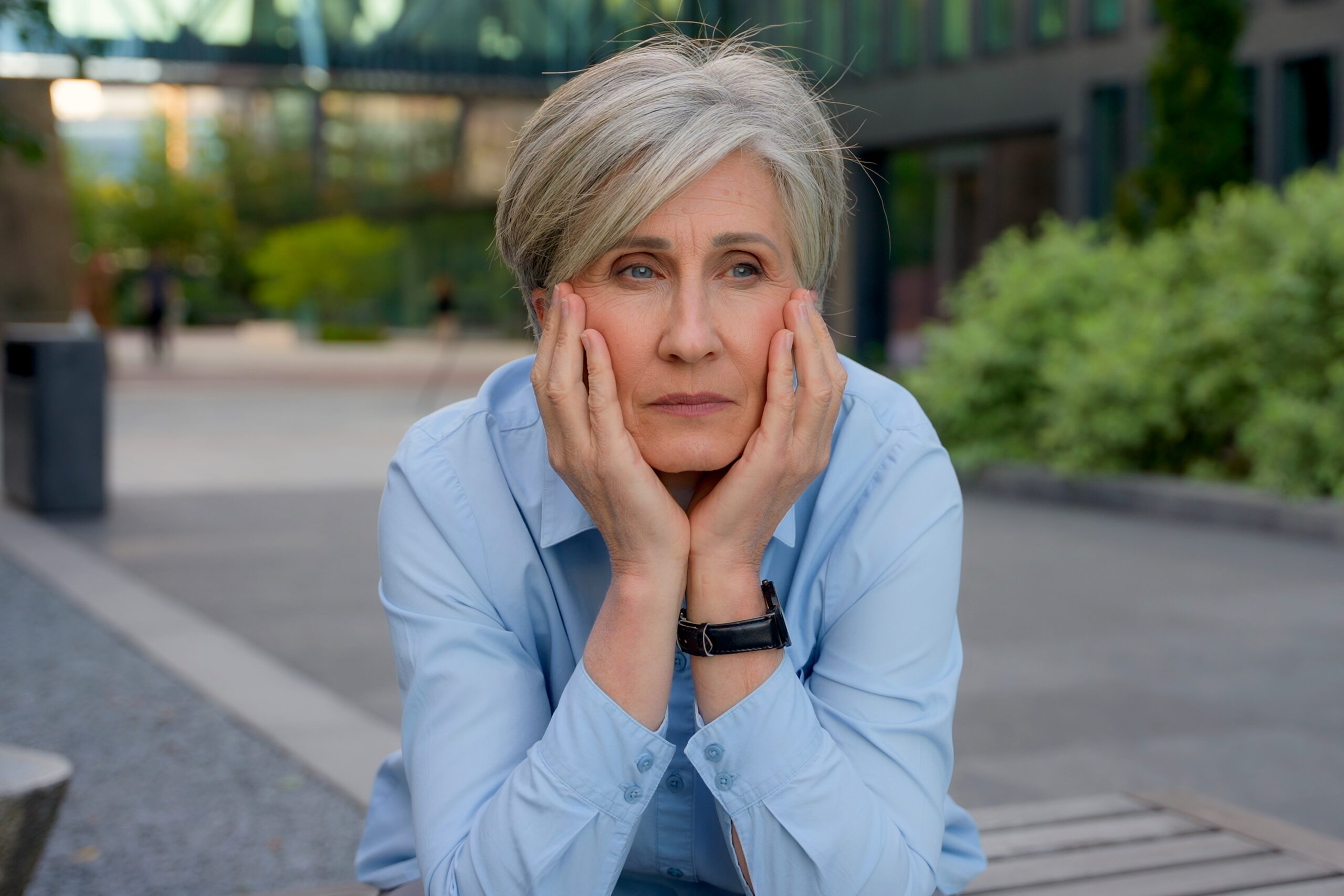 A woman with short gray hair, wearing a light blue shirt and a black wristwatch, rests her head in her hands while sitting outdoors. The background features a modern building, greenery, and a pathway. She appears thoughtful or contemplative.