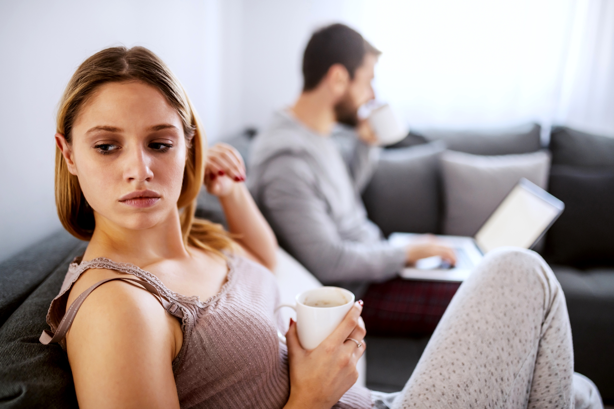 A woman with blond hair sits on a couch, holding a mug and looking over her shoulder with a concerned expression. A bearded man in the background sits on the same couch, focused on his laptop while drinking from a mug. Both are wearing comfortable clothes.