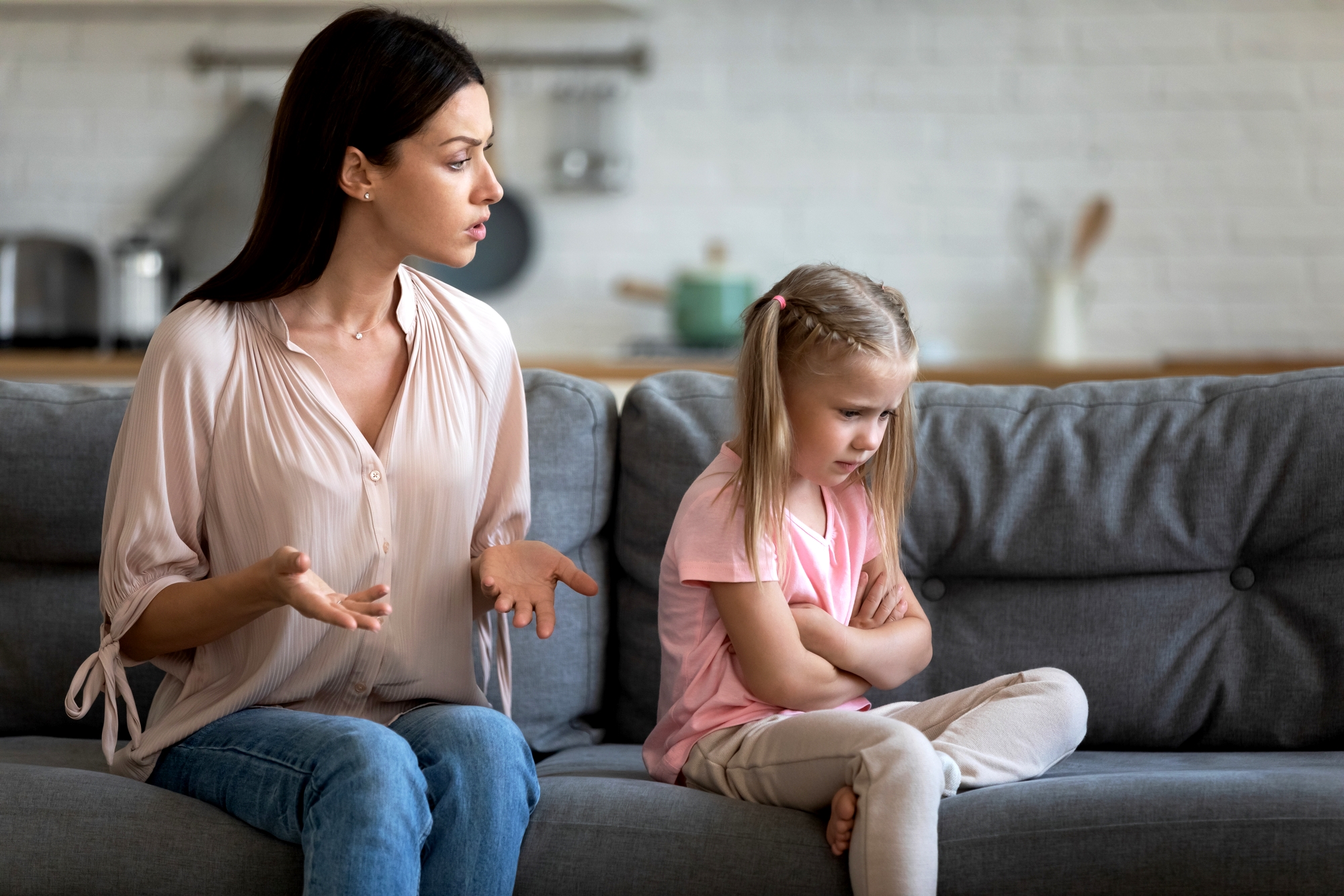 A woman with dark hair in a light blouse sits on a gray sofa, gesturing as she talks to a young girl with blond pigtails in a pink shirt who has her arms crossed and looks upset. The background shows a kitchen out of focus.