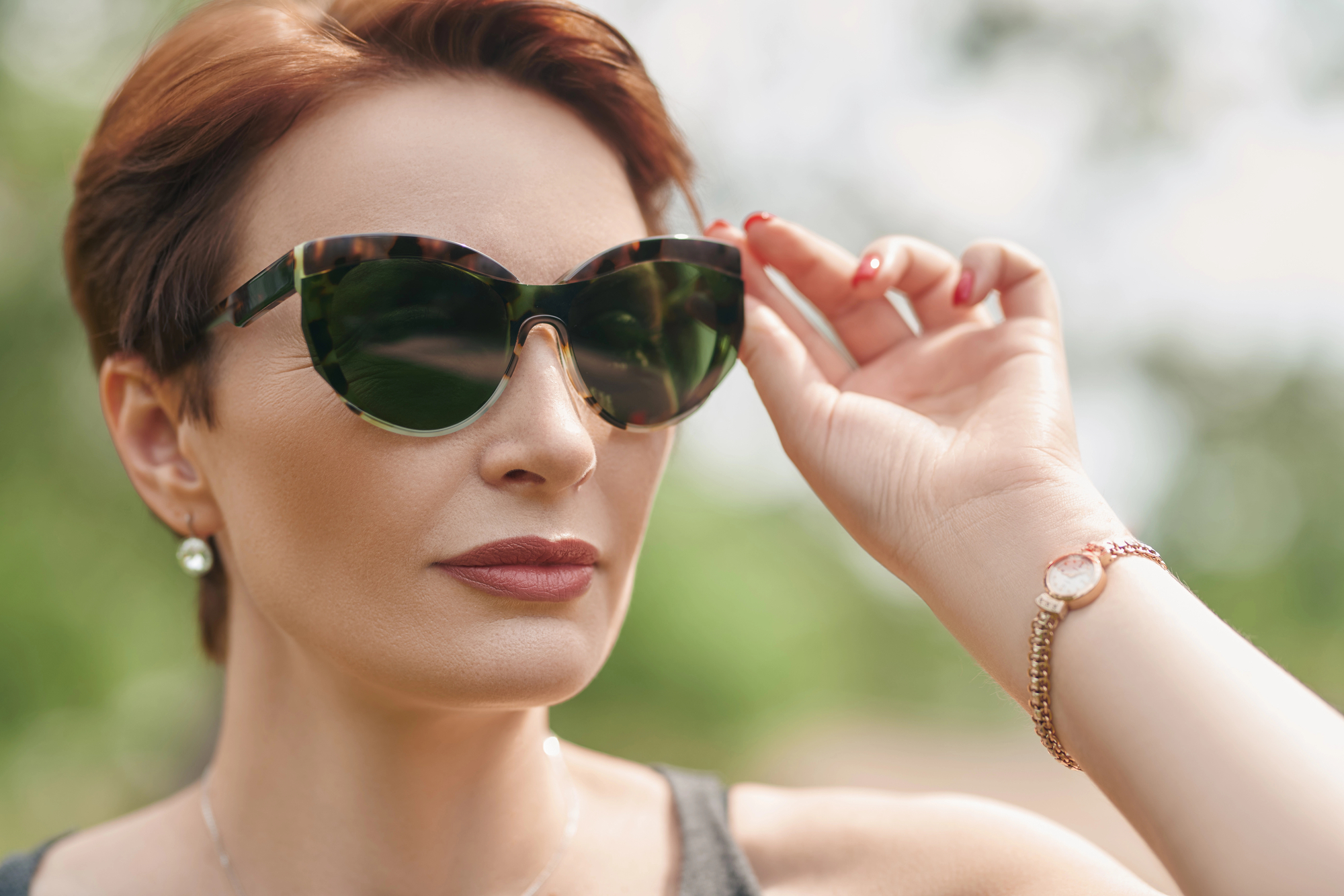 A woman with short auburn hair is wearing large cat-eye sunglasses and a sleeveless top. She is touching the frame of her sunglasses with her left hand, showcasing a watch on her wrist. The background is blurred and appears to be a green outdoor setting.
